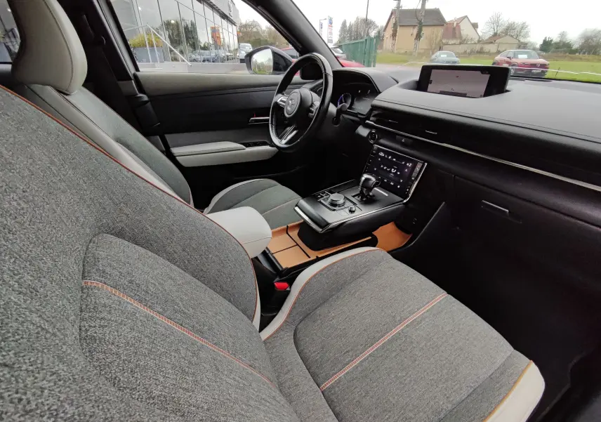 Intérieur du Mazda MX-30 E-SKYACTIV 145 Makoto 2023, vue côté conducteur, sièges gris clair avec surpiqûres orange et console centrale moderne.