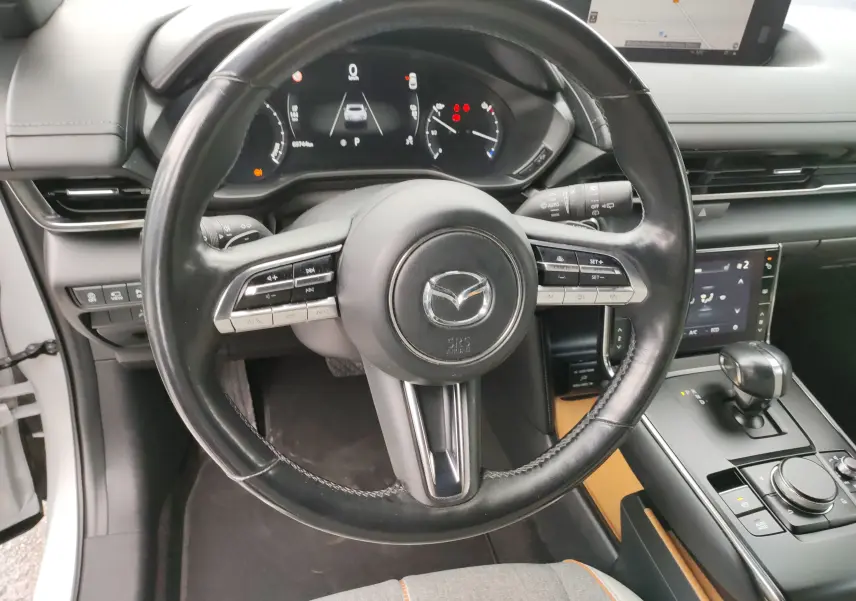 Vue intérieure centrée sur le volant noir de la Mazda MX-30 gris clair 2023 avec tableau de bord digital et console moderne.