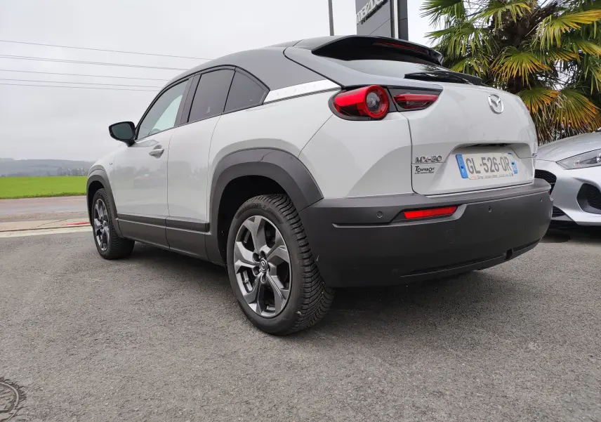 Vue 3/4 arrière droite d'une Mazda MX-30 électrique gris clair avec toit noir et jantes alliage, stationnée en extérieur.