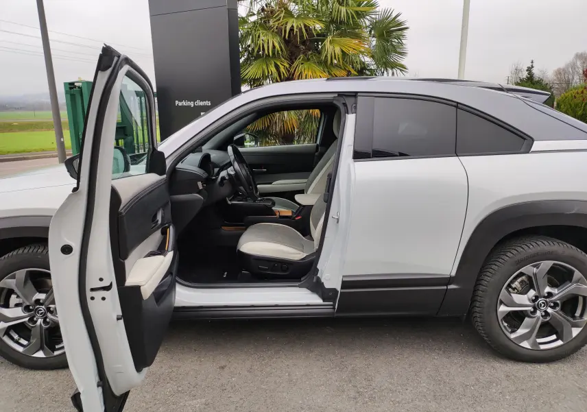 Vue latérale gauche du Mazda MX-30 gris clair 2023 avec porte avant ouverte dévoilant l’intérieur clair et moderne.