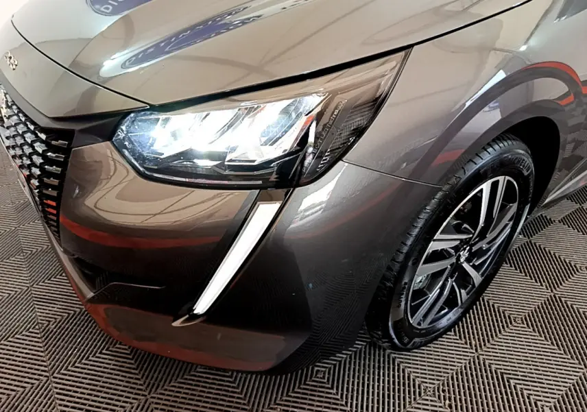 Vue 3/4 avant droit d'une Peugeot 208 gris foncé avec phares LED allumés et jante alliage visible.