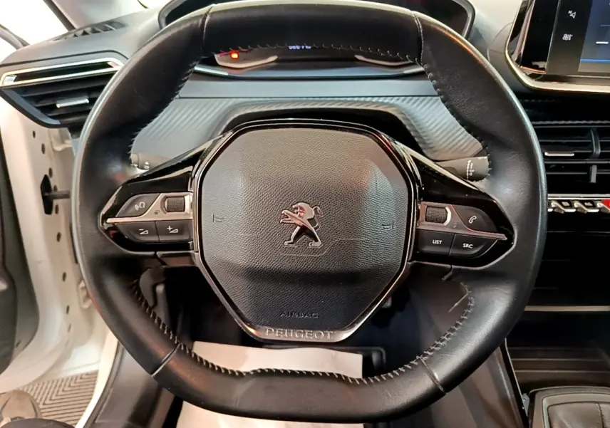 Vue rapprochée du volant noir de la Peugeot 208 blanc 2021, avec commandes intégrées et logo lion central.