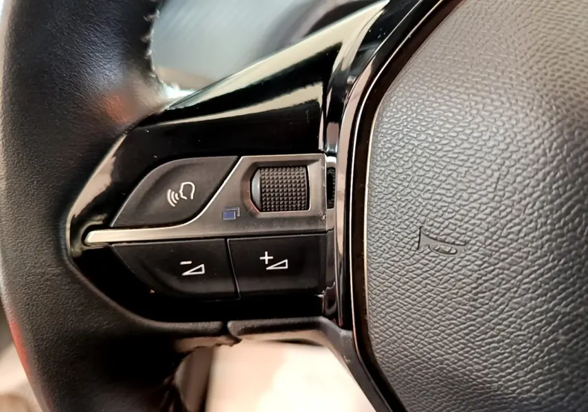Gros plan sur les commandes gauche du volant noir de la Peugeot 208 blanche, avec boutons de volume et appel vocal.