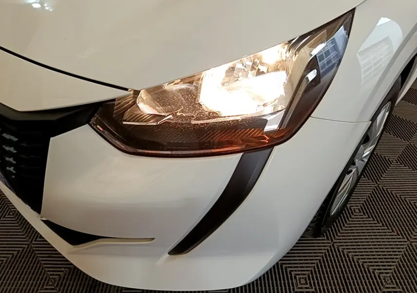 Gros plan sur l'avant droit blanc de la Peugeot 208 2021, mettant en valeur le phare allumé et la calandre noire.