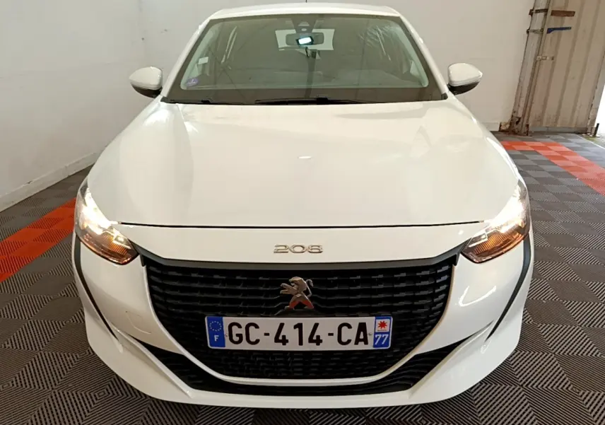 Vue avant d'une Peugeot 208 blanche avec calandre noire et phares allumés dans un garage.