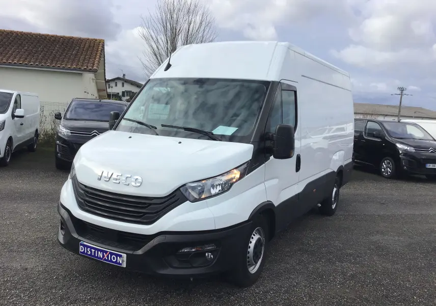 Fourgon Iveco Daily blanc vu en 3/4 avant droit sur un parking, avec calandre noire et phares allumés.