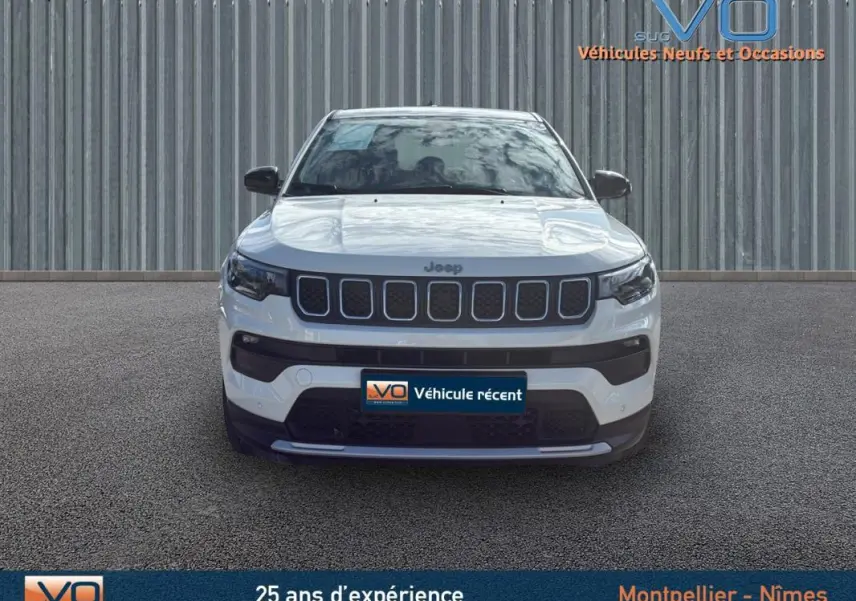 Vue frontale d'un Jeep Compass blanc 2024 avec calandre noire et phares LED, sur fond industriel gris.