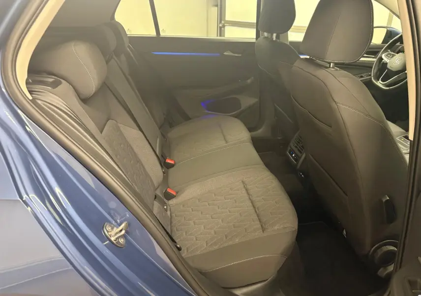 Vue intérieure arrière côté gauche de la Volkswagen Golf 1.5 TSI 2025 en Bleu Anémone, sièges tissu gris et éclairage d'ambiance bleu.