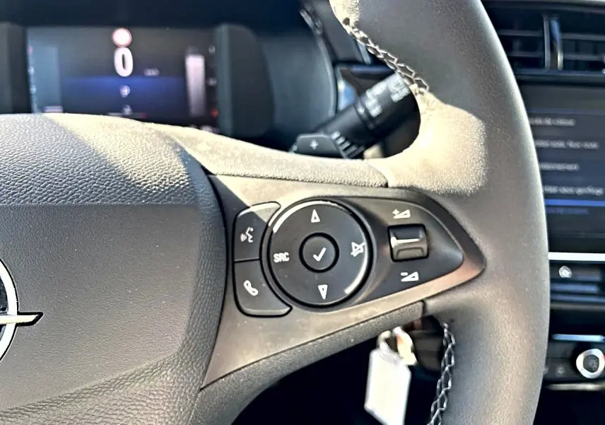 Gros plan sur le volant multifonction noir de l’Opel Corsa gris alu, avec commandes audio et affichage numérique en arrière-plan.