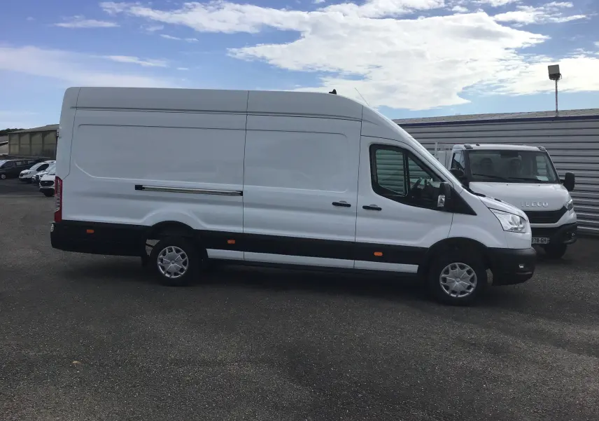 Fourgon Ford Transit blanc vu de profil côté gauche sur un parking avec un ciel partiellement nuageux.