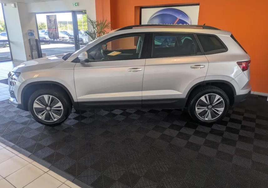 Vue de profil côté gauche d’un Skoda Karoq gris argent 2023, SUV compact exposé en intérieur sur sol noir.