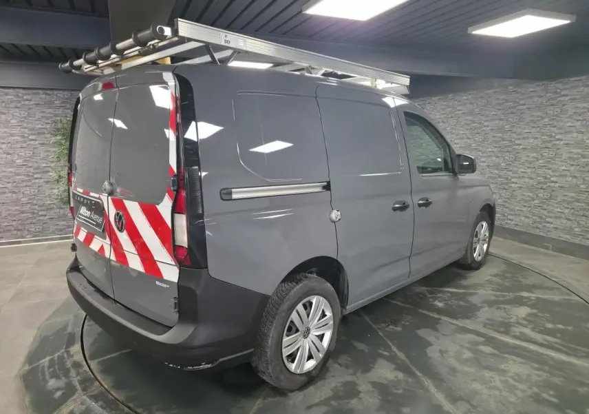 Vue 3/4 arrière droite du Volkswagen Caddy Cargo gris avec galerie de toit et marquage rouge et blanc sur portes arrière.