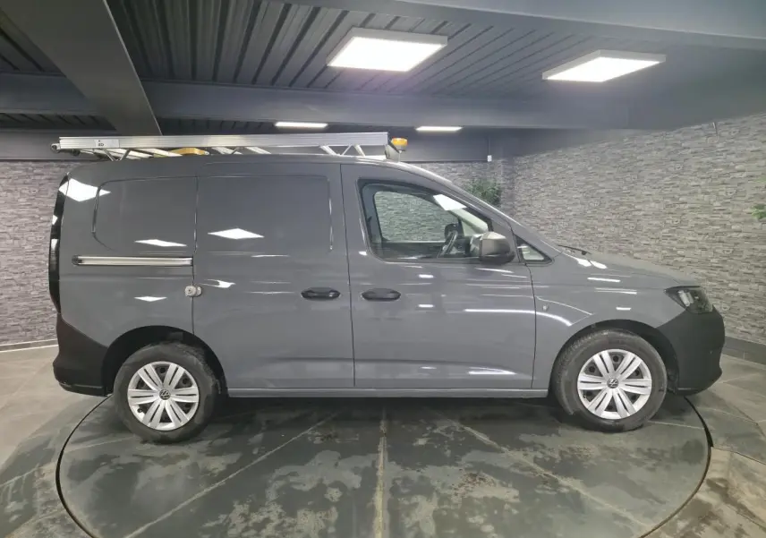 Vue de profil droit du Volkswagen Caddy Cargo gris avec galerie de toit et jantes acier, dans un showroom intérieur.