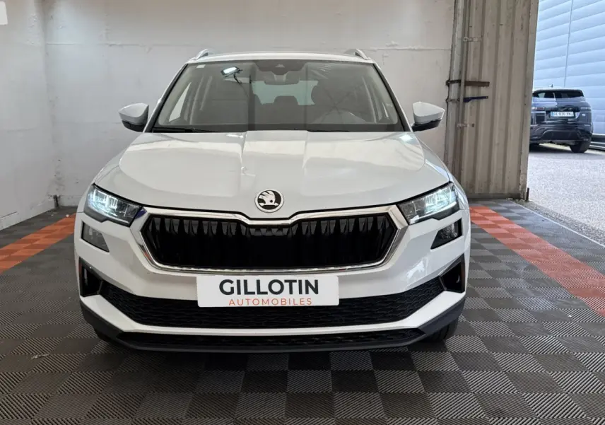 Vue de face du SUV blanc SKODA KAROQ 2024 avec calandre noire et phares LED allumés dans un garage.