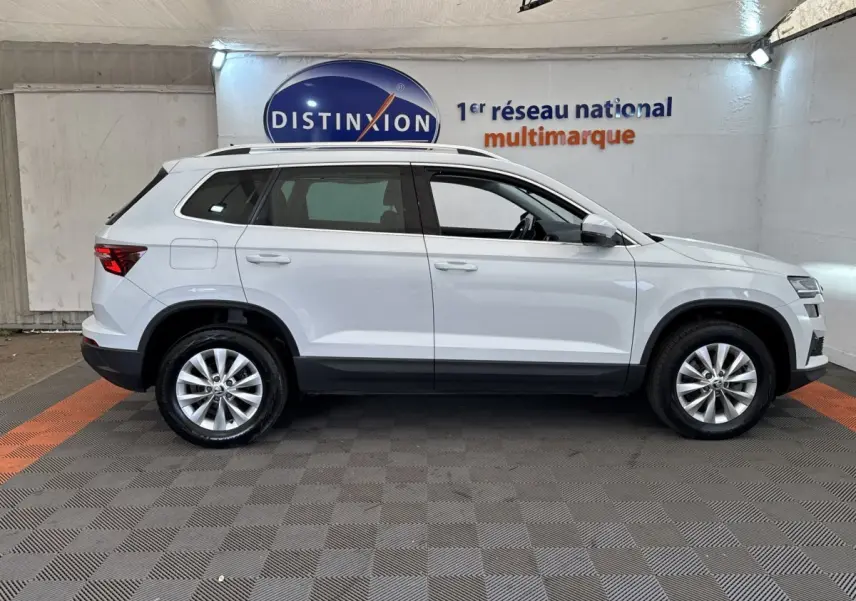 Vue latérale droite d'un Skoda Karoq blanc 2024 avec jantes alliage et barres de toit noires dans un showroom.