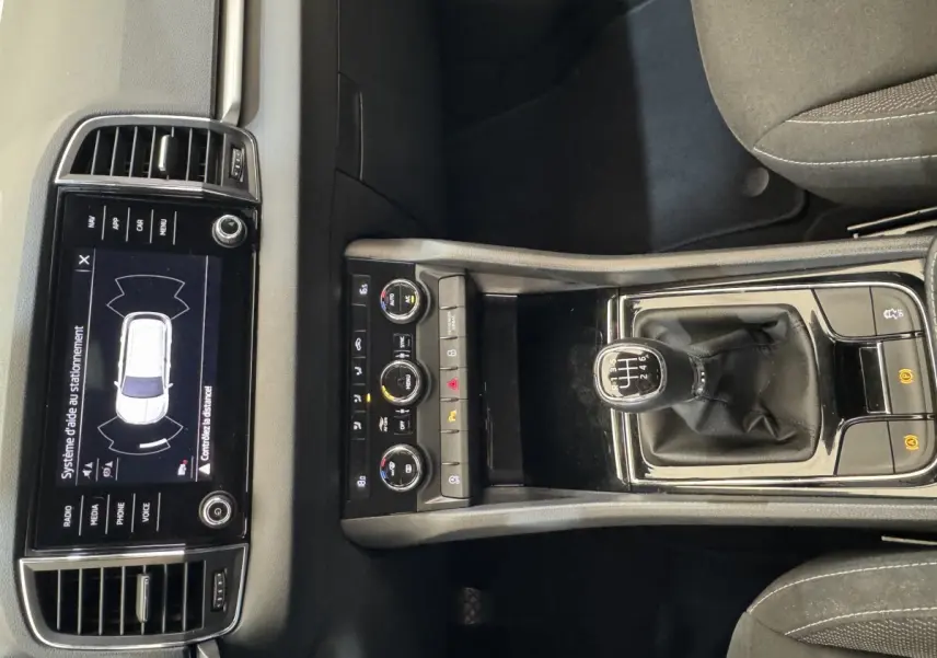 Vue intérieure centrée sur la console centrale du Skoda Karoq blanc, avec levier de vitesses manuel et écran tactile affichant l'aide au stationnement.