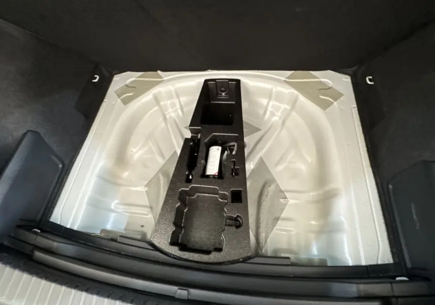 Vue plongeante sur le coffre ouvert d’un Skoda Karoq blanc 2024, montrant le rangement pour roue de secours et outils.