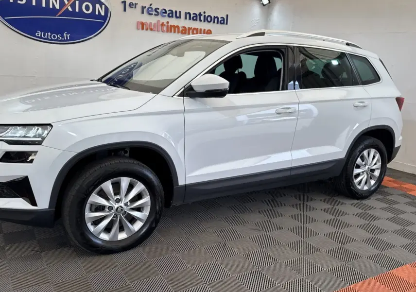 Vue de profil côté gauche d'un Skoda Karoq blanc 2024 avec jantes alliage et barres de toit noires en showroom.