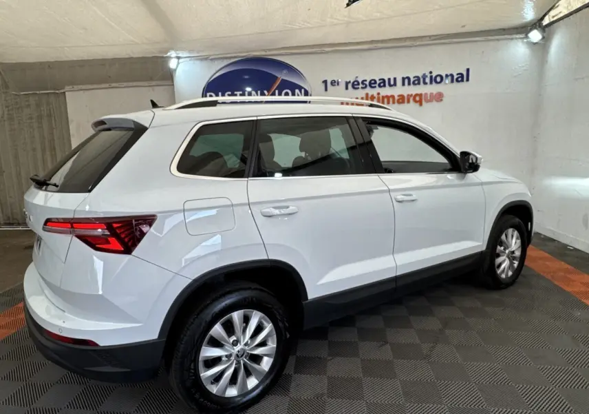 Vue 3/4 arrière droite d’un Skoda Karoq blanc 2024 dans un showroom, avec feux arrière LED et barres de toit noires.