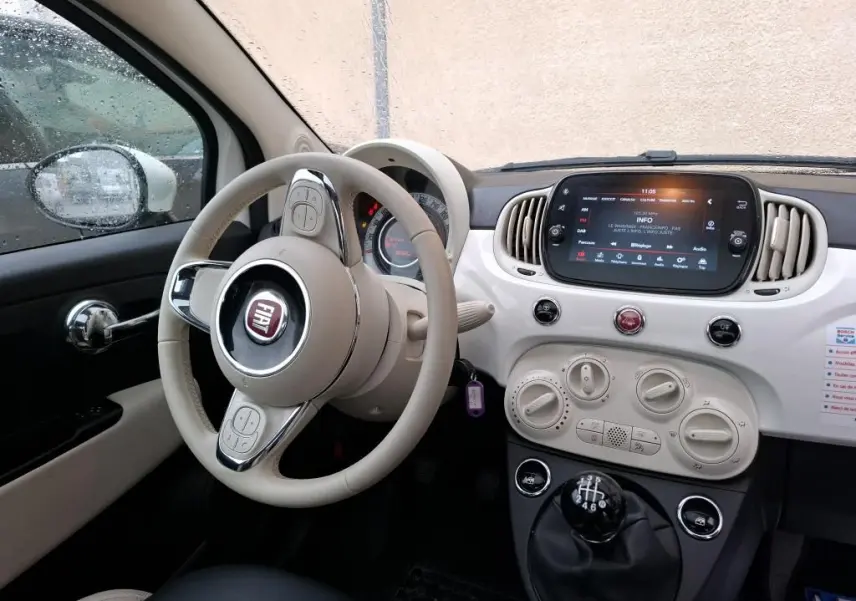 Vue intérieure côté conducteur du tableau de bord blanc et beige d'une FIAT 500 2022 avec volant beige et levier de vitesses manuel noir.