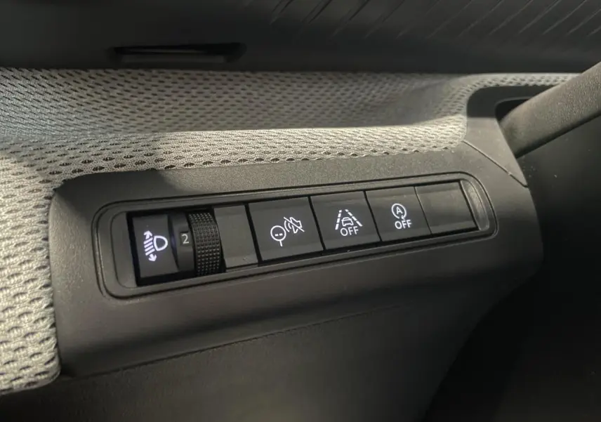 Gros plan sur les commandes d’assistance et réglage des phares à gauche du tableau de bord d’une Citroën C3 gris foncé.