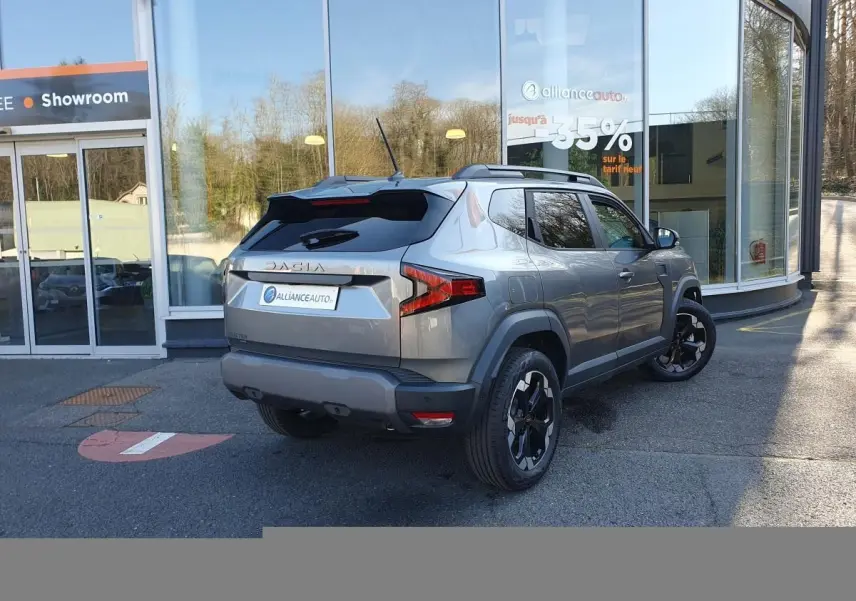 Vue 3/4 arrière droite du Dacia Duster 2026 gris schiste avec jantes alliage noires et toit noir, devant un showroom.