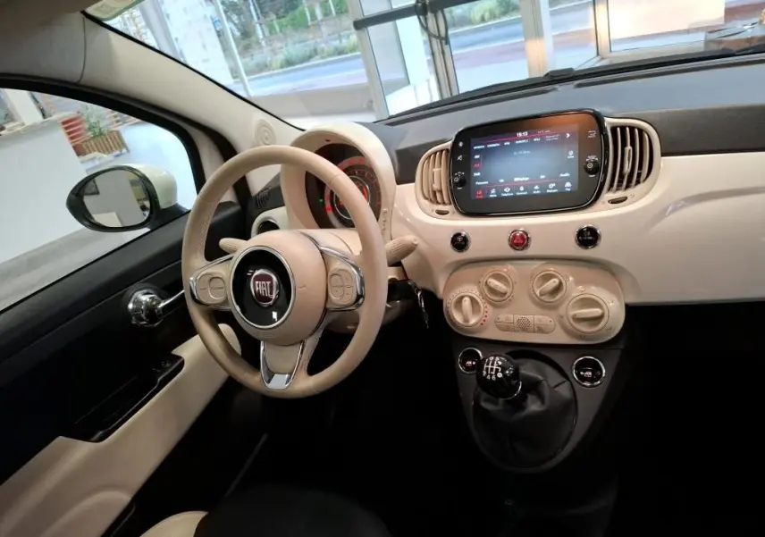 Vue intérieure côté conducteur de la FIAT 500 blanche avec tableau de bord beige et levier de vitesses manuel visible.