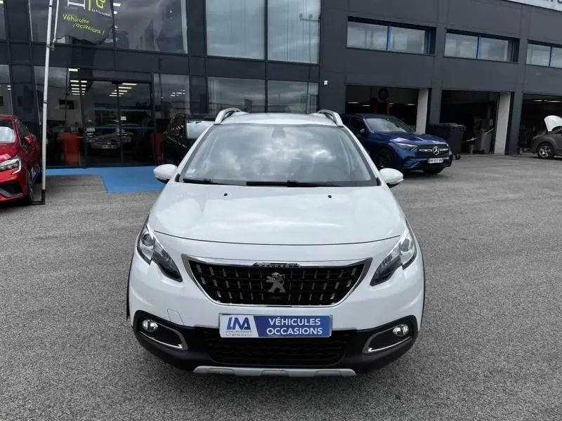Peugeot 2008 blanc vue de face, avec calandre noire et badge lion, stationnée devant un garage de véhicules d'occasion.