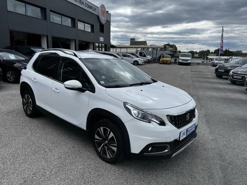 Peugeot 2008 blanc vue 3/4 avant droit, avec jantes alliage et barres de toit, stationnée devant un bâtiment commercial.