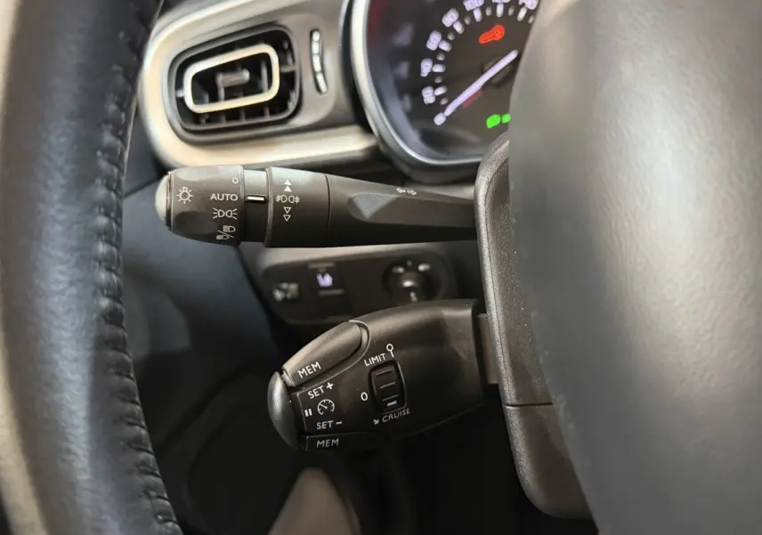 Gros plan sur les commandes au volant noir et tableau de bord éclairé d'une Citroën C3 gris clair, côté gauche.