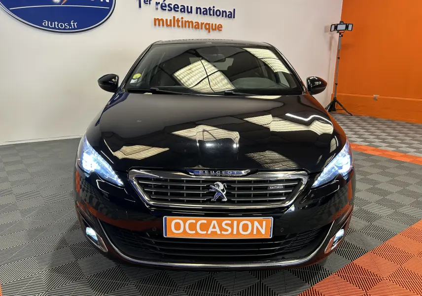 Peugeot 308 noir vue de face avec phares allumés et plaque orange "OCCASION" en intérieur showroom.