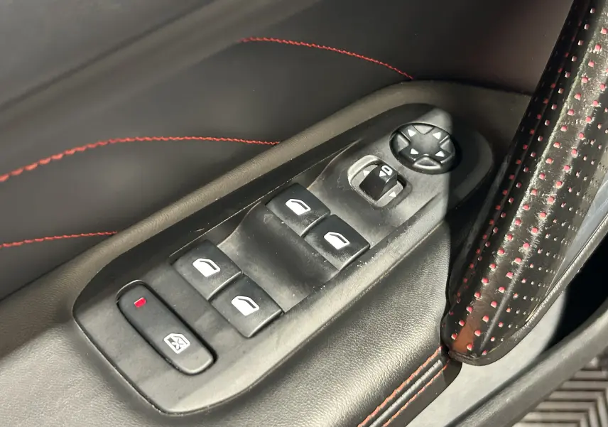 Gros plan sur les commandes de lève-vitres et réglage des rétroviseurs côté conducteur, avec surpiqûres rouges sur cuir noir.