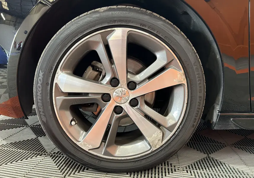 Gros plan sur la roue avant gauche d'une Peugeot 308 noire, jante alliage 5 branches avec traces d'usure visibles.