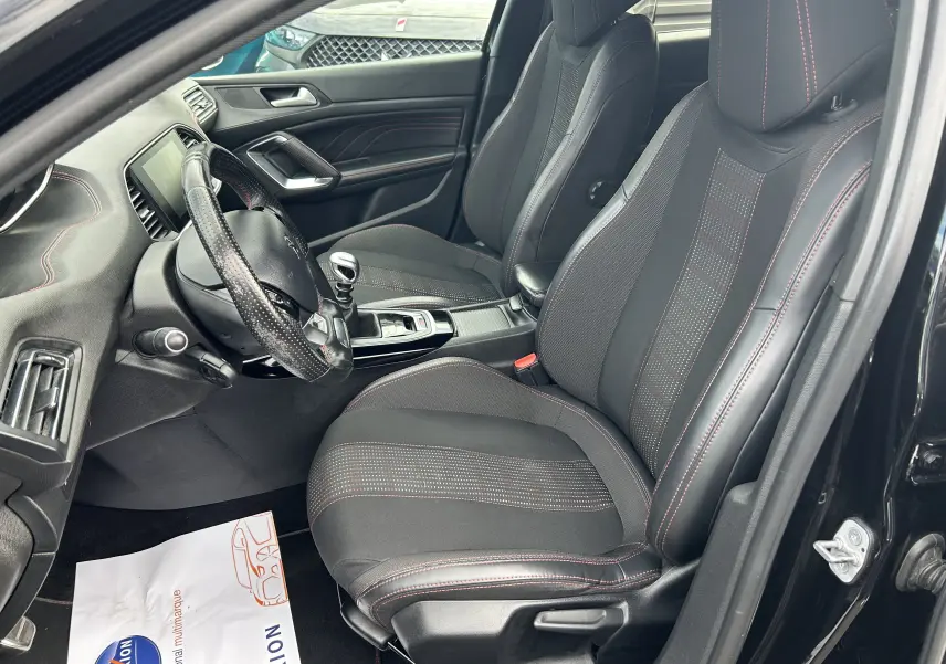 Vue intérieure côté conducteur de la Peugeot 308 noire, avec sièges sport noirs et coutures rouges visibles.