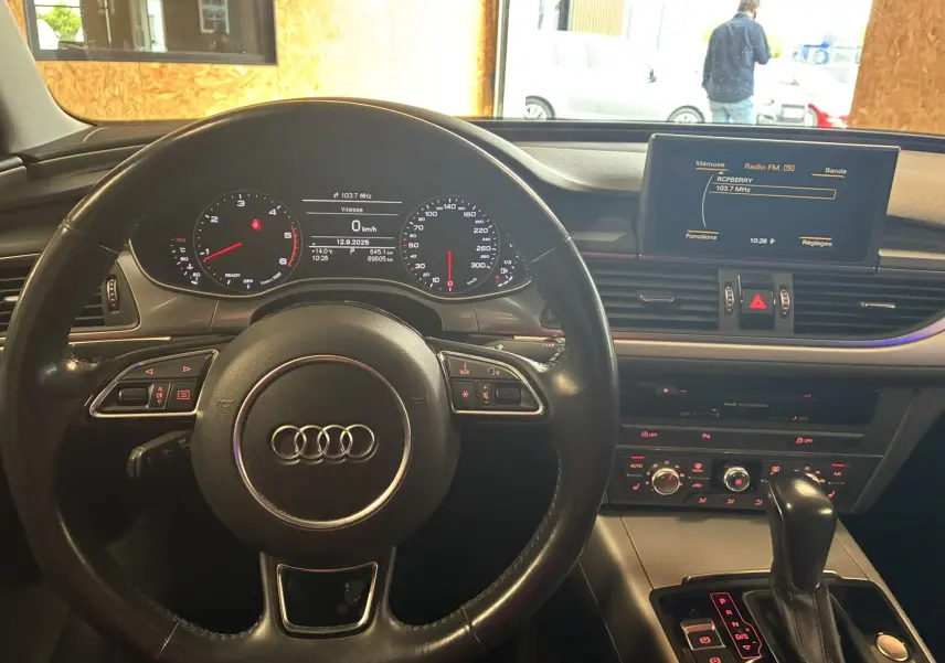 Vue intérieure centrée sur le volant noir et le tableau de bord digital d'une Audi A6 Avant 2017, avec écran multimédia allumé.