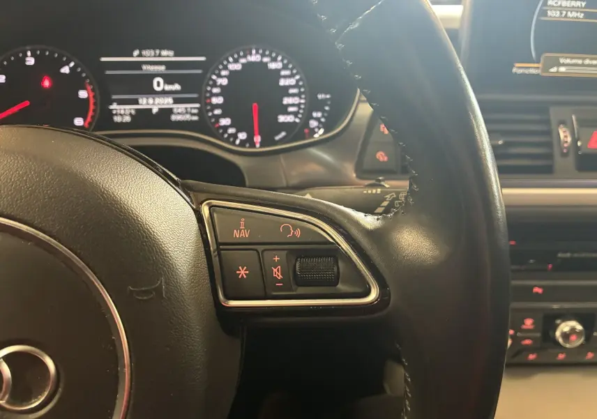 Gros plan sur le volant noir de l'Audi A6 Avant 2017, avec commandes audio et tableau de bord lumineux en arrière-plan.