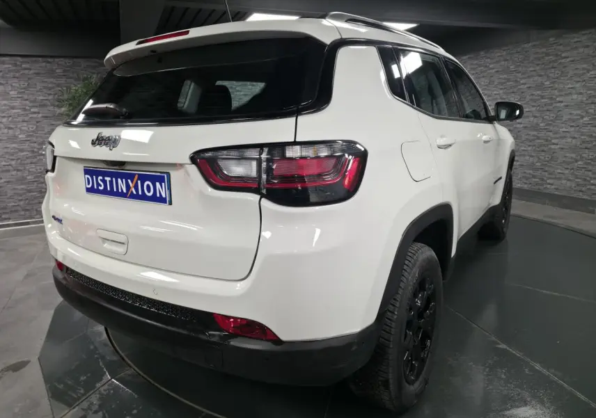 Vue 3/4 arrière droite du Jeep Compass blanc avec jantes noires et logo distinctif sur fond intérieur gris.