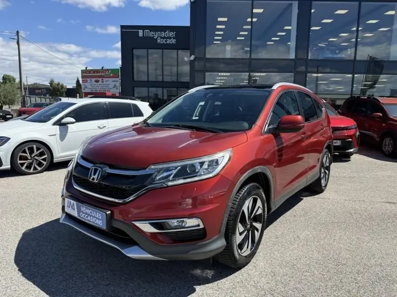 Honda CR-V rouge métal en 3/4 avant droit, avec jantes alliage et calandre noire brillante, stationné devant un bâtiment vitré.
