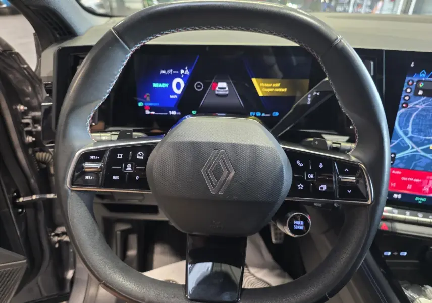 Vue rapprochée du volant en textile noir avec insert Alcantara et logo Renault, tableau de bord numérique en arrière-plan.