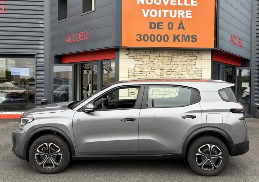 Vue de profil côté gauche d'une Citroën C3 Aircross électrique gris foncé garée devant un bâtiment avec panneau orange.