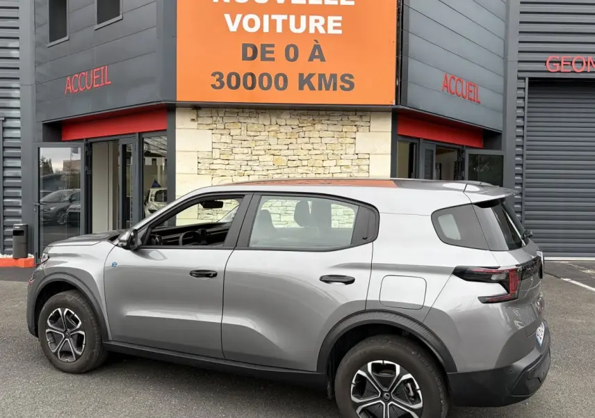 Vue de profil côté gauche d'un Citroën C3 Aircross électrique gris foncé garé devant un bâtiment avec panneau orange.