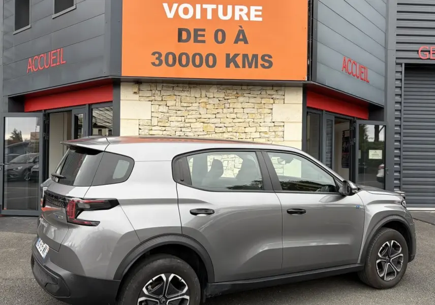 Vue latérale droite d'un Citroën C3 Aircross électrique gris foncé garé devant un bâtiment avec enseigne orange.
