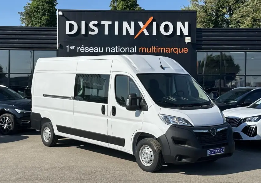 Fourgon blanc Opel Movano L3H2 en 3/4 avant droit, avec porte latérale vitrée et crochet d'attelage visible.