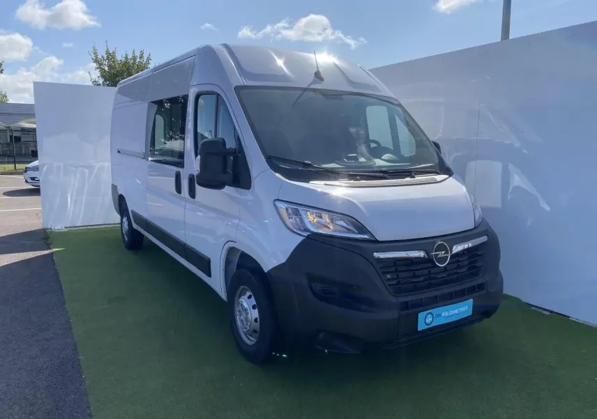 Vue 3/4 avant gauche d'un Opel Movano Fourgon blanc avec porte latérale vitrée et calandre noire.
