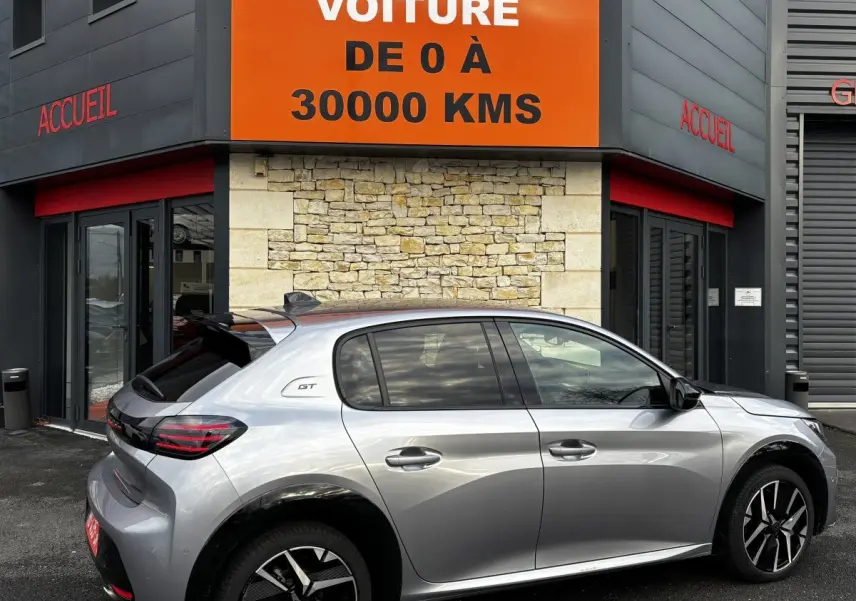 Peugeot 208 Hybrid 110 e-DCS6 GT gris clair vue de profil côté droit devant un bâtiment avec panneau orange "votre nouvelle voiture".