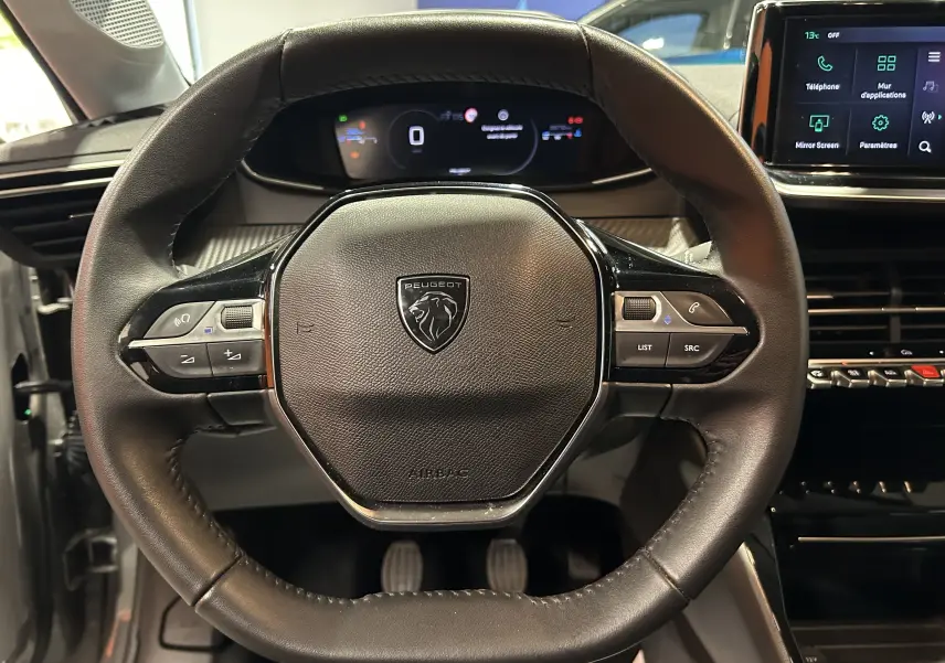 Vue centrée sur le volant cuir noir de la Peugeot 208 2025 avec tableau de bord numérique et écran tactile à droite.