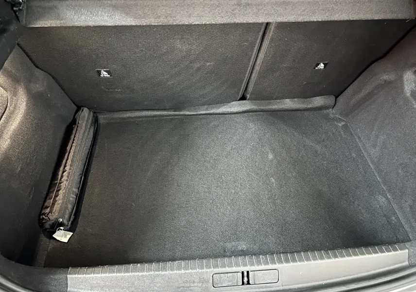 Coffre vide de Peugeot 208 II Turbo 100 Allure, vue plongeante montrant le tapis noir et une trousse rangée sur le côté.