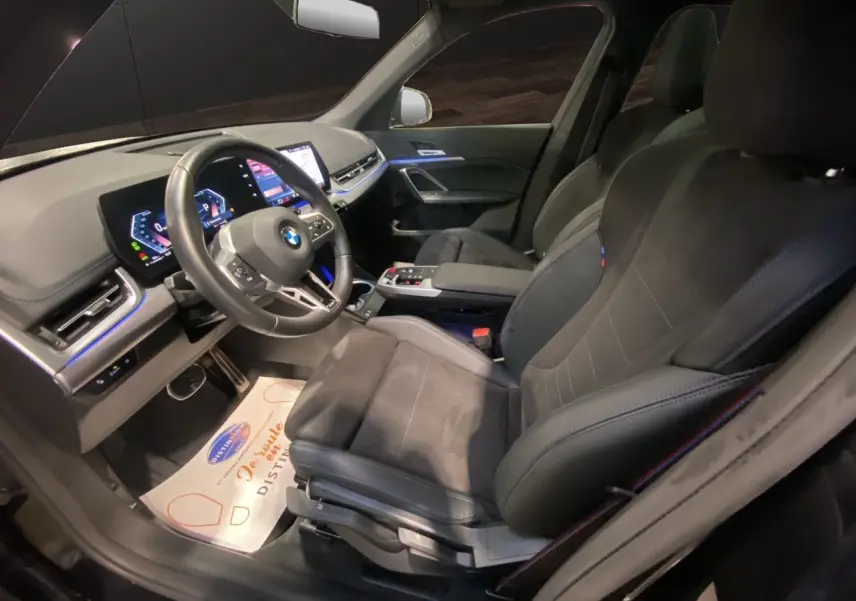 Intérieur BMW X1 2025 vu côté conducteur, sièges noirs M Sport avec tableau de bord digital et éclairage d'ambiance bleu.
