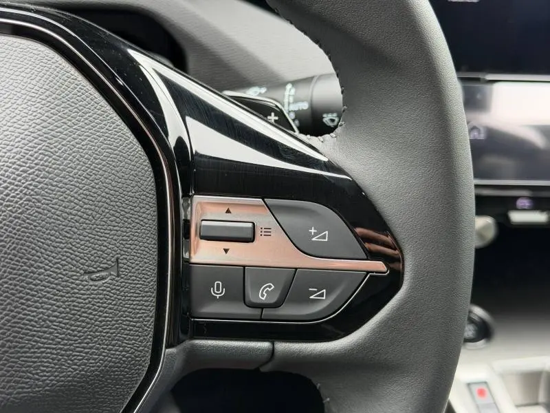 Gros plan sur les commandes multifonctions côté gauche du volant noir du Peugeot 308 blanc Okénite hybride 2025.