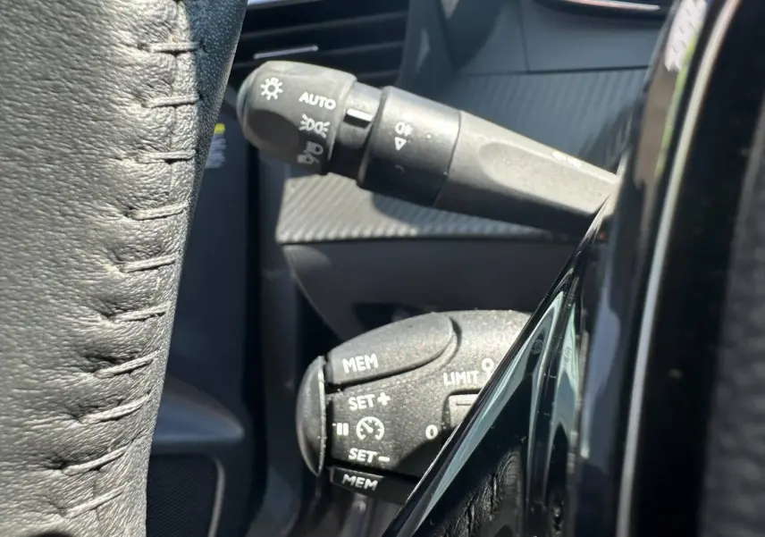 Gros plan sur les commandes au volant noir de la Peugeot 208 1.2 100ch Allure, avec détails des boutons de régulateur et phares.